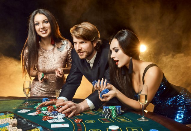 Two beautiful women and young man play on poker table in casino, focus on man and brunette. Passion, cards, chips, alcohol, dice, gambling, casino - it is entertainment. Dangerous fun card game for money. Smoke background.