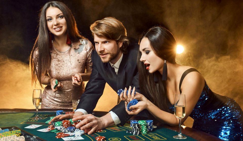Two beautiful women and young man play on poker table in casino, focus on man and brunette. Passion, cards, chips, alcohol, dice, gambling, casino - it is entertainment. Dangerous fun card game for money. Smoke background.
